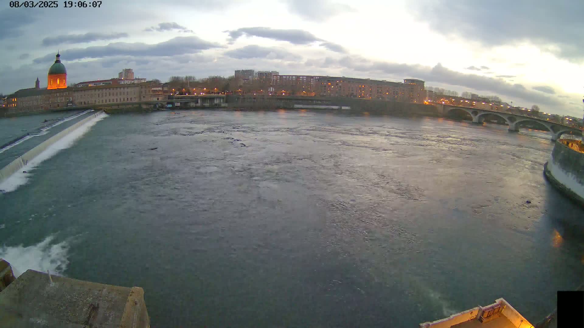 A wide shot of a river flowing past a city under a cloudy sky at dusk, with a dam visible in the foreground and a bridge in the background.