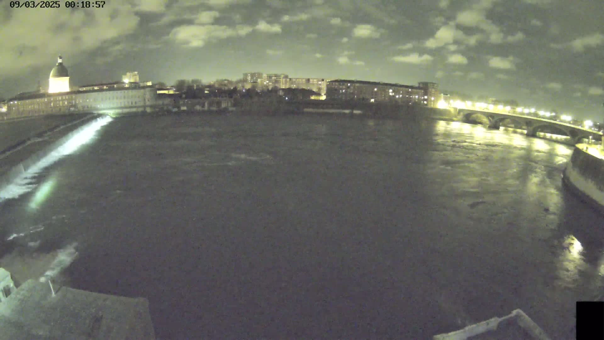 A nighttime view of a river flowing past city buildings and a bridge under a cloudy sky.