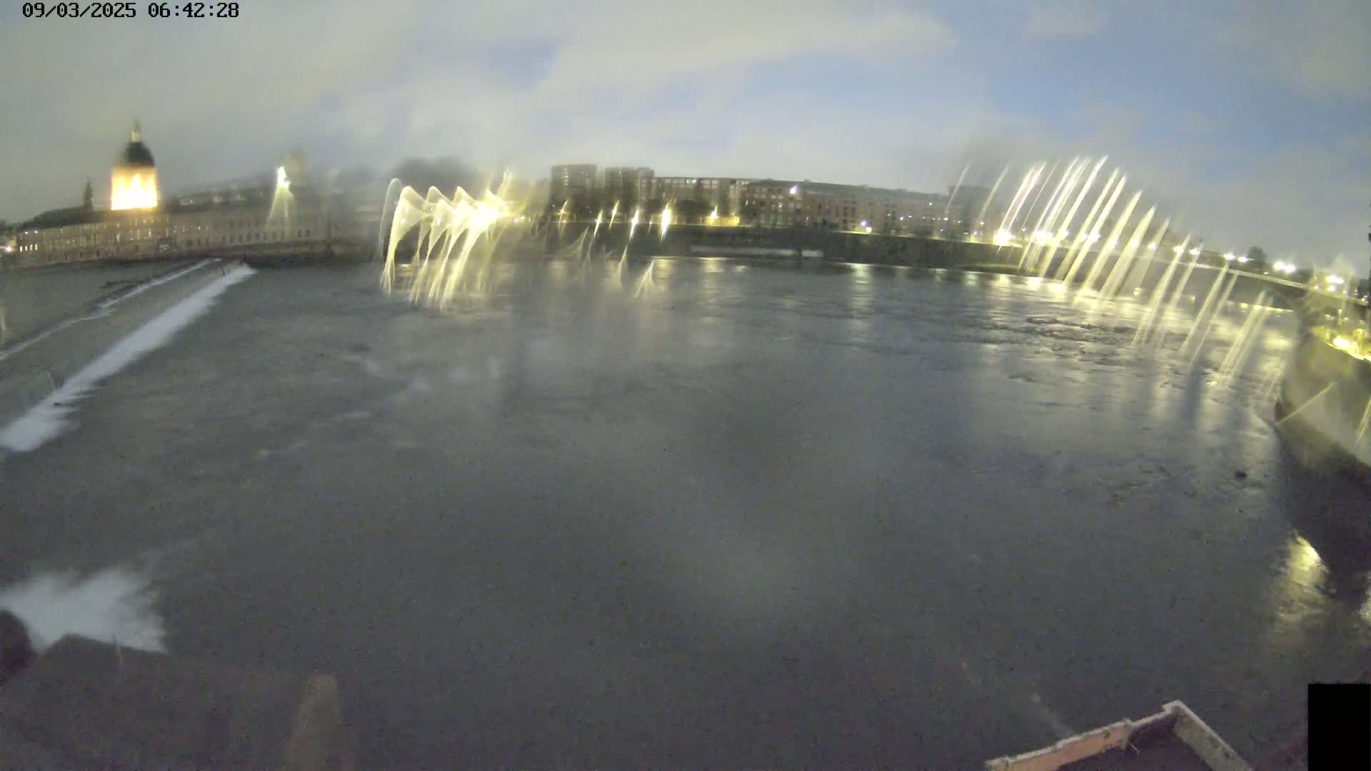 A nighttime view of a river with illuminated fountains in front of city buildings under a cloudy sky.