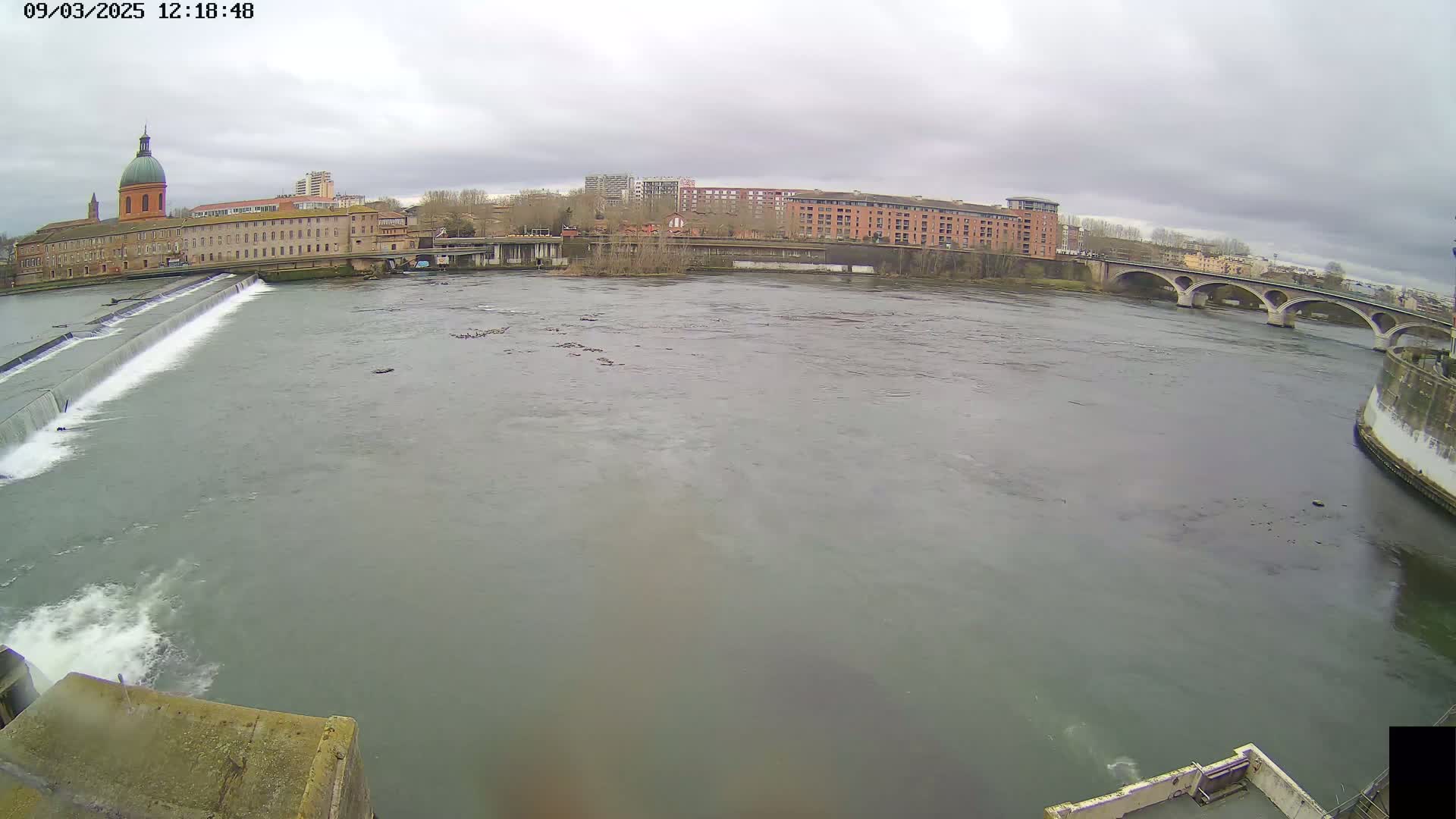 A wide shot shows a river flowing under a bridge, past a building with a dome, on an overcast day.