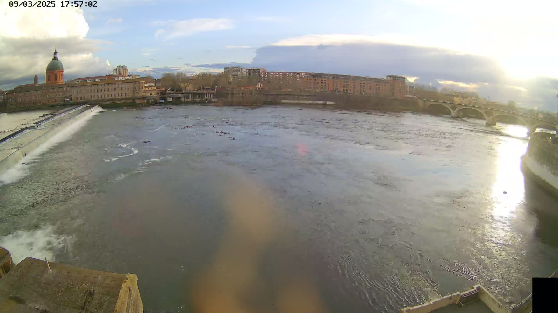A wide shot of a river flowing past a city, under a partly cloudy sky, with a dam and bridge visible.