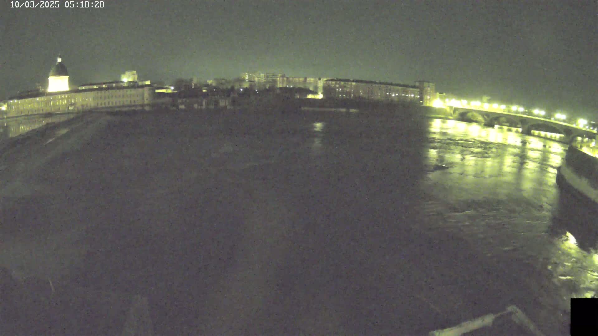 A nighttime view shows a river flowing past a city with buildings lit up along the banks and a bridge illuminated across the water.