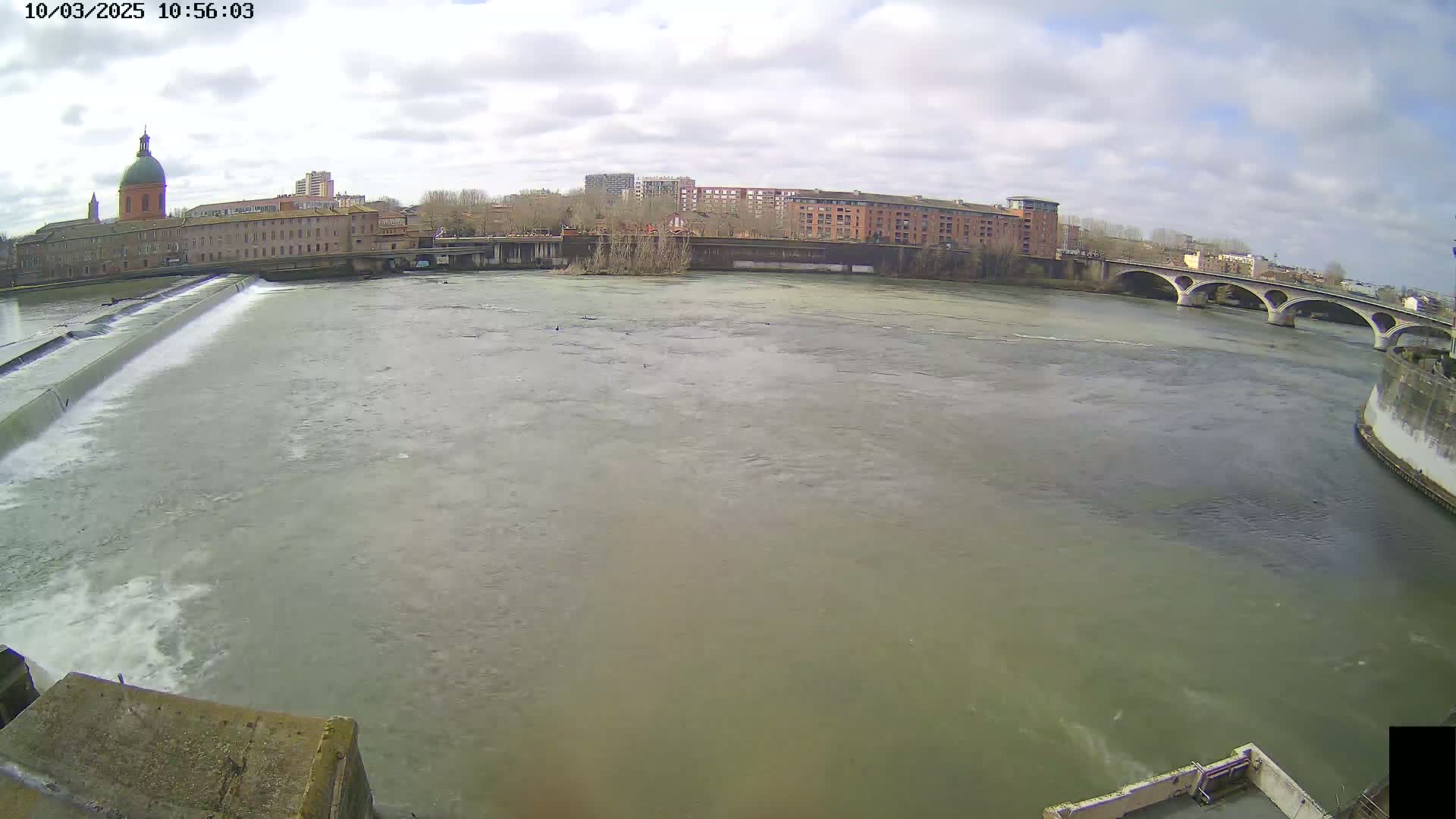 A wide shot of a river flowing under a bridge on a partly cloudy day, with a dam and city buildings in the background.