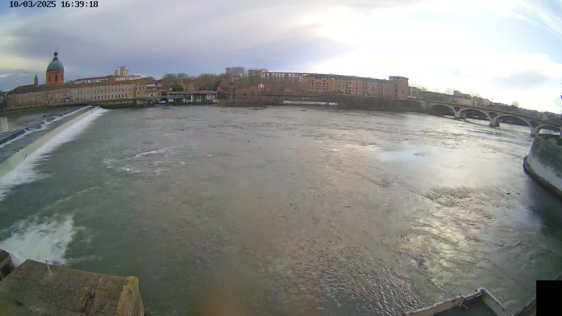 A wide shot of a river flowing past a city with buildings and a bridge under an overcast sky.