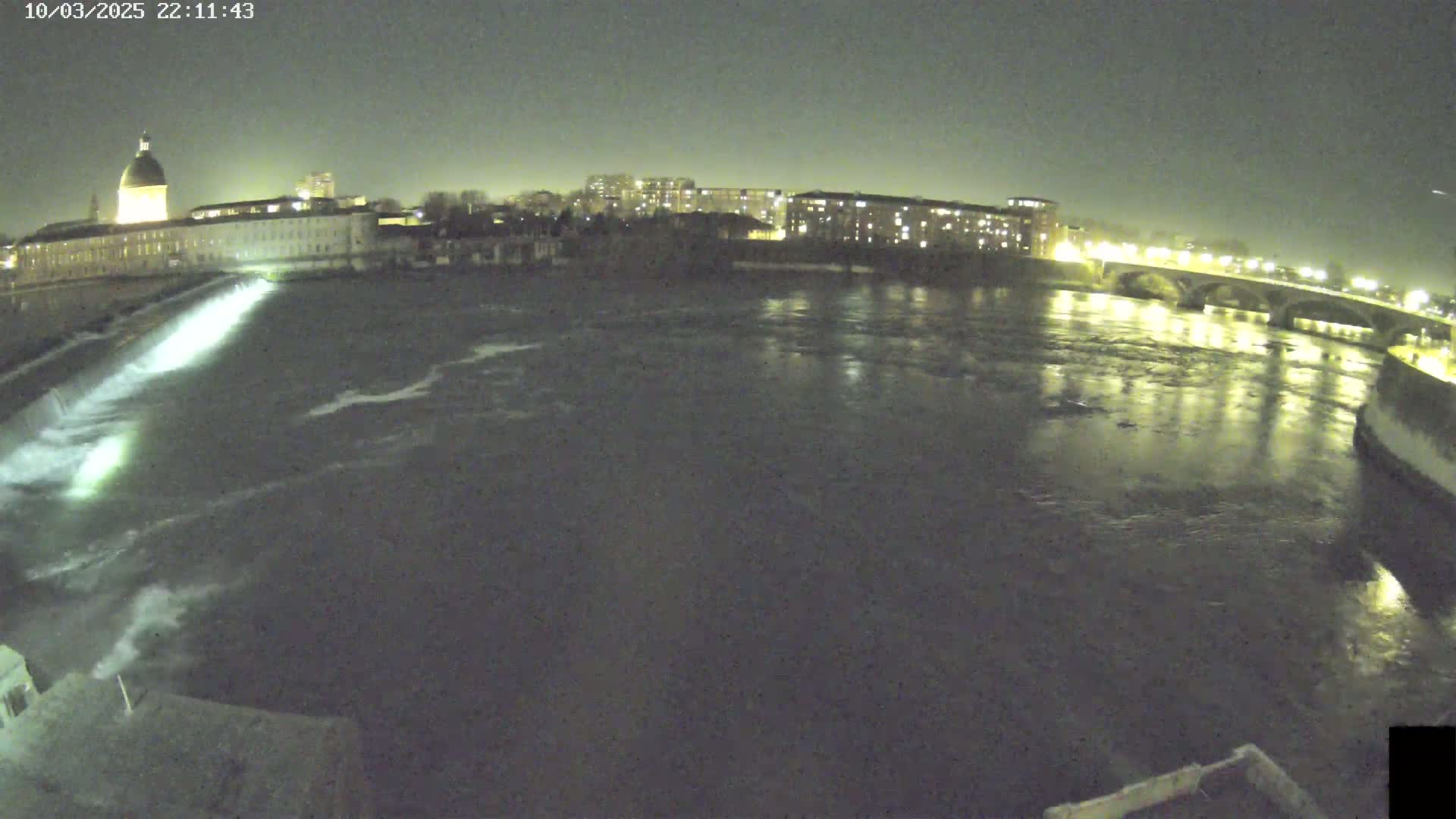 A nighttime view of a river flowing past a city, with a dam visible in the foreground and lights illuminating buildings and a bridge in the background.