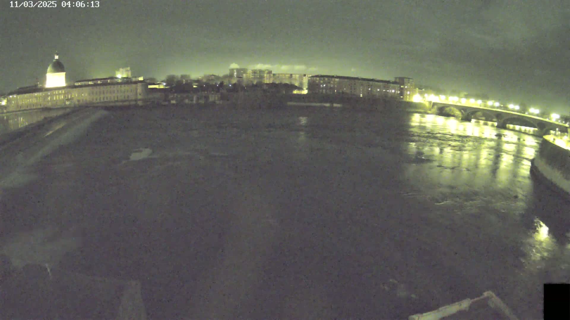 A wide nighttime shot shows a river flowing under a bridge past buildings with lights on, under a dark, cloudy sky.