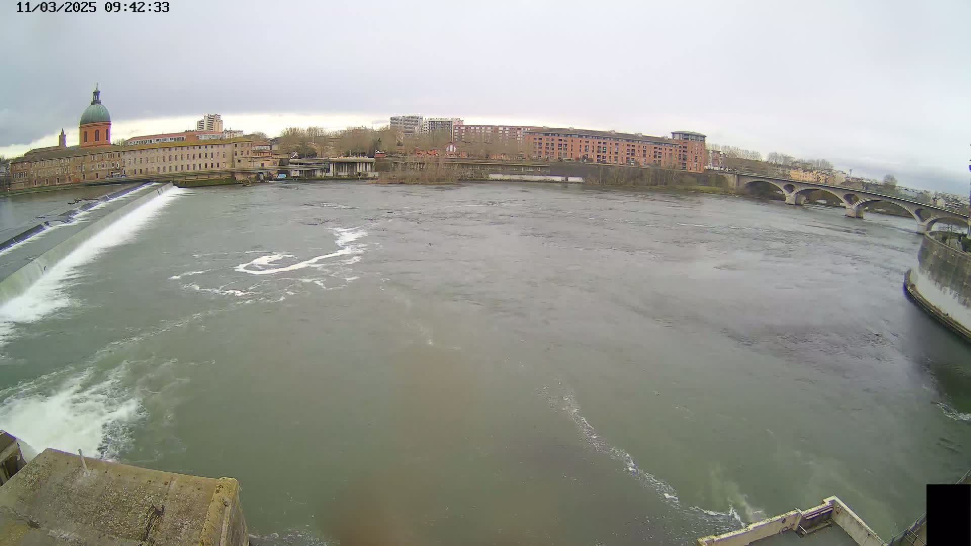 A wide, murky river flows past a city under an overcast sky, featuring a dam in the foreground and a stone bridge in the background.