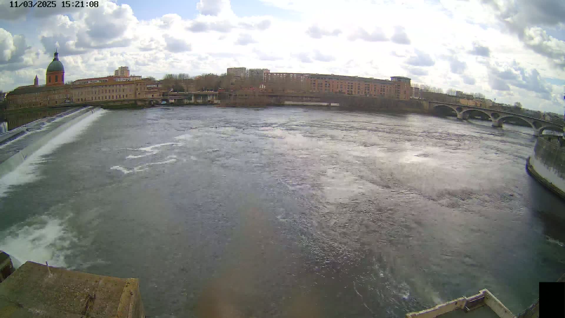 A wide shot of a river flowing under a bridge on a partly cloudy day, with a city and buildings visible in the background.