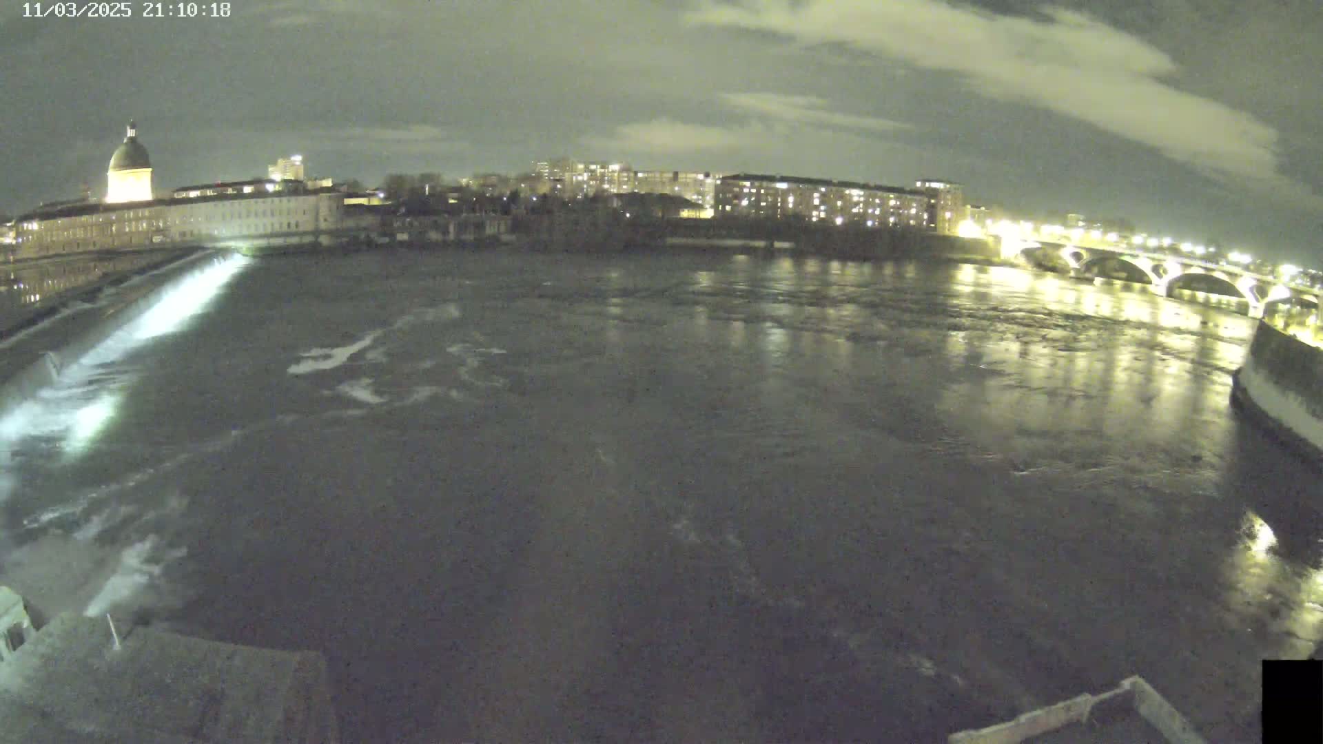 A nighttime view of a river flowing past a city with a building featuring a dome, illuminated by city lights under a cloudy sky.