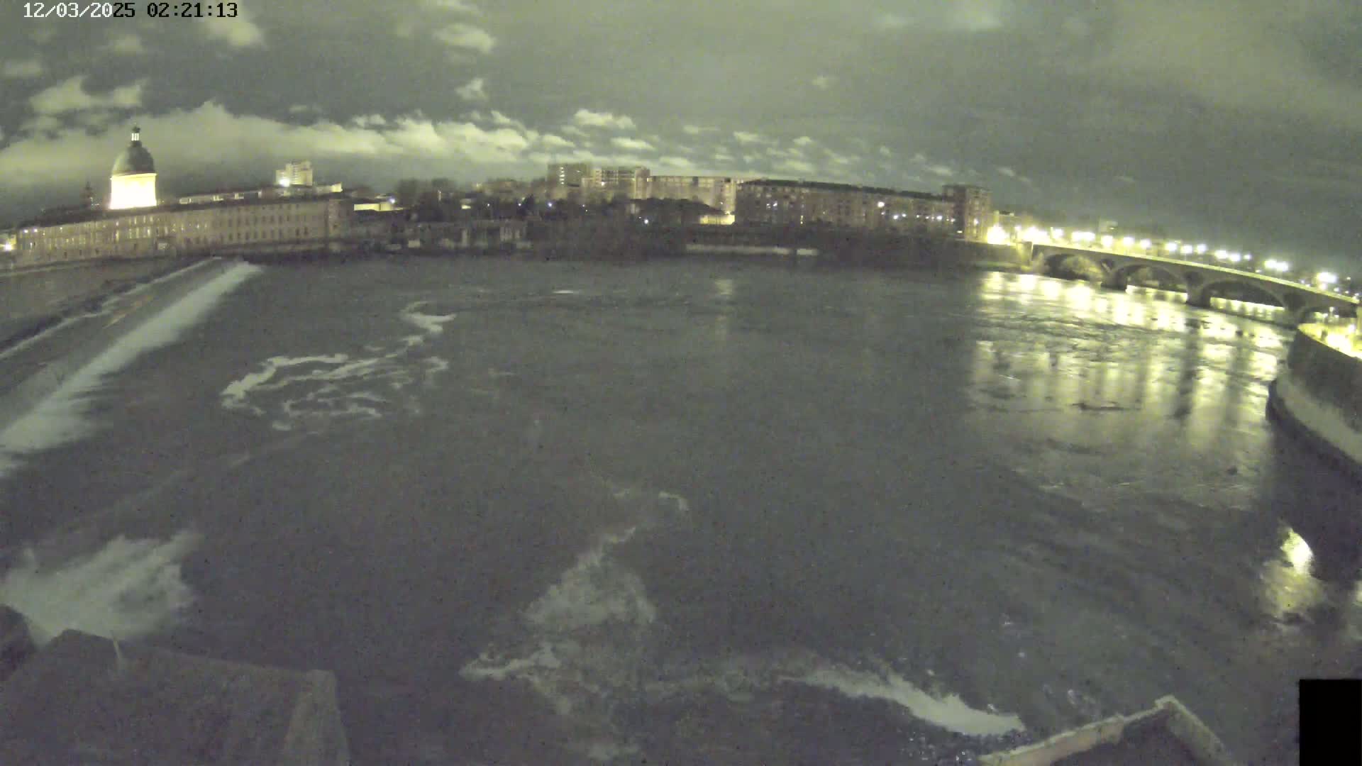 A nighttime view of a river flowing past a city with buildings and a bridge under a partly cloudy sky.