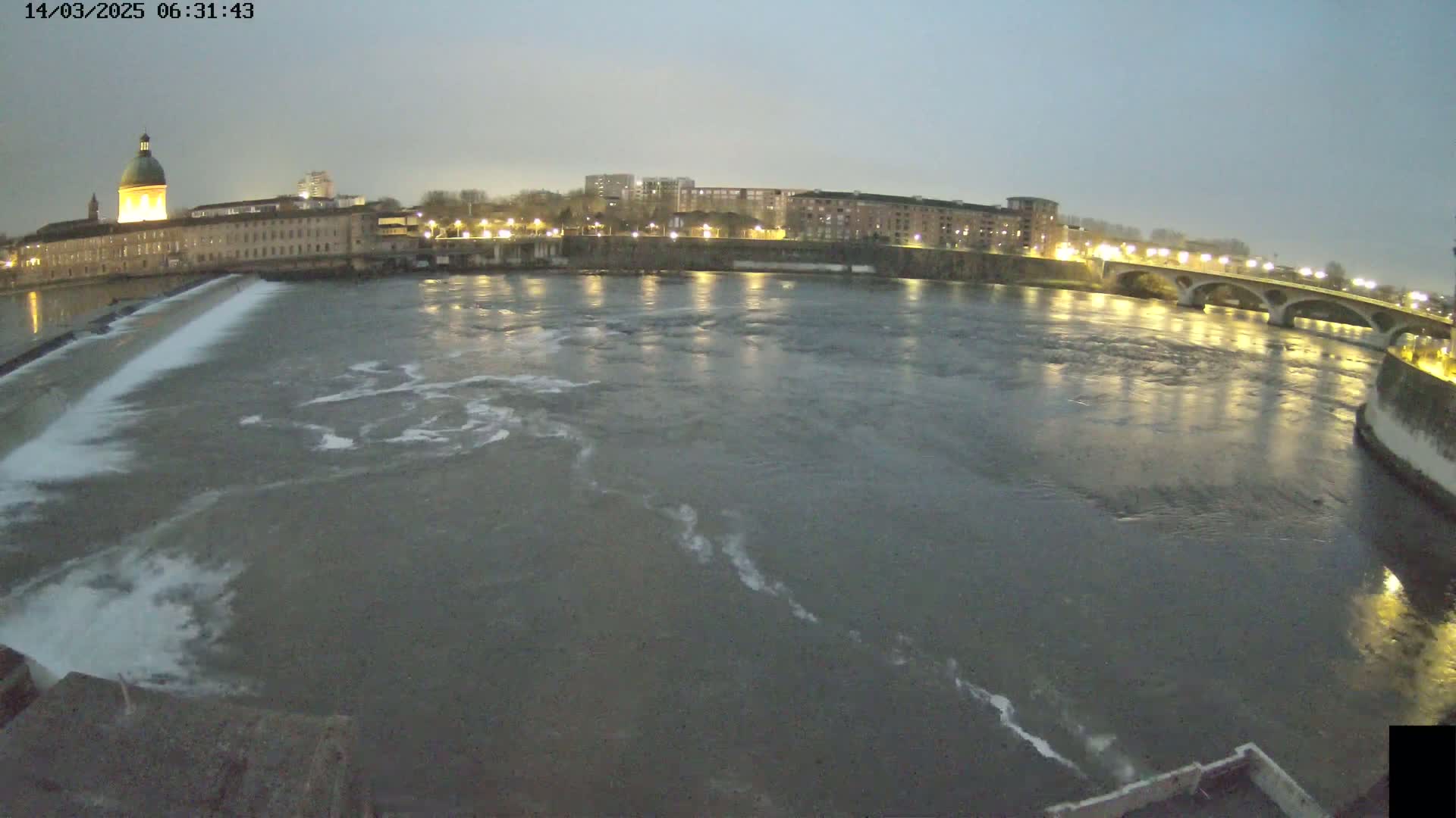 A wide shot at twilight shows a river flowing past a city with a dam and bridge, under a dusky sky.