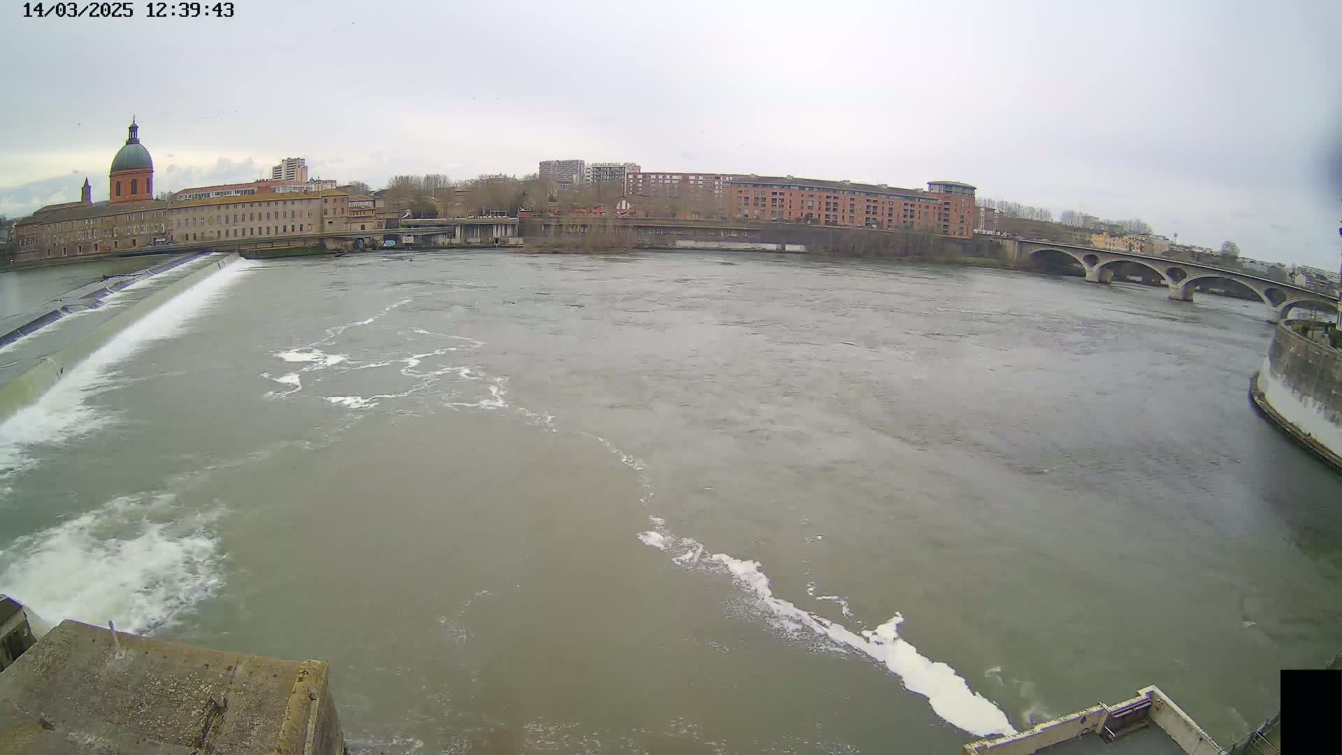 A wide, murky river flows past a dam and under a bridge, with buildings and overcast skies in the background.