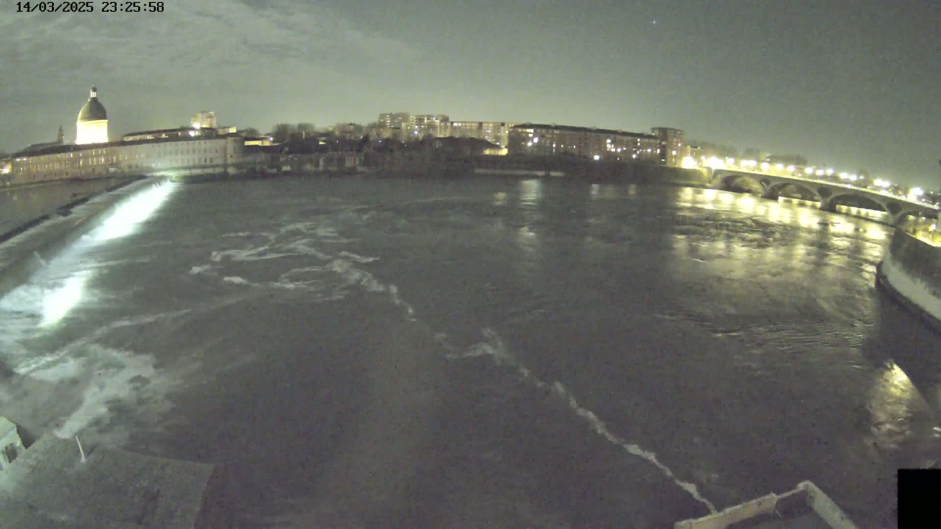 A nighttime view of a river flowing past a city, with a dam visible in the foreground and a bridge in the distance under a mostly clear sky.