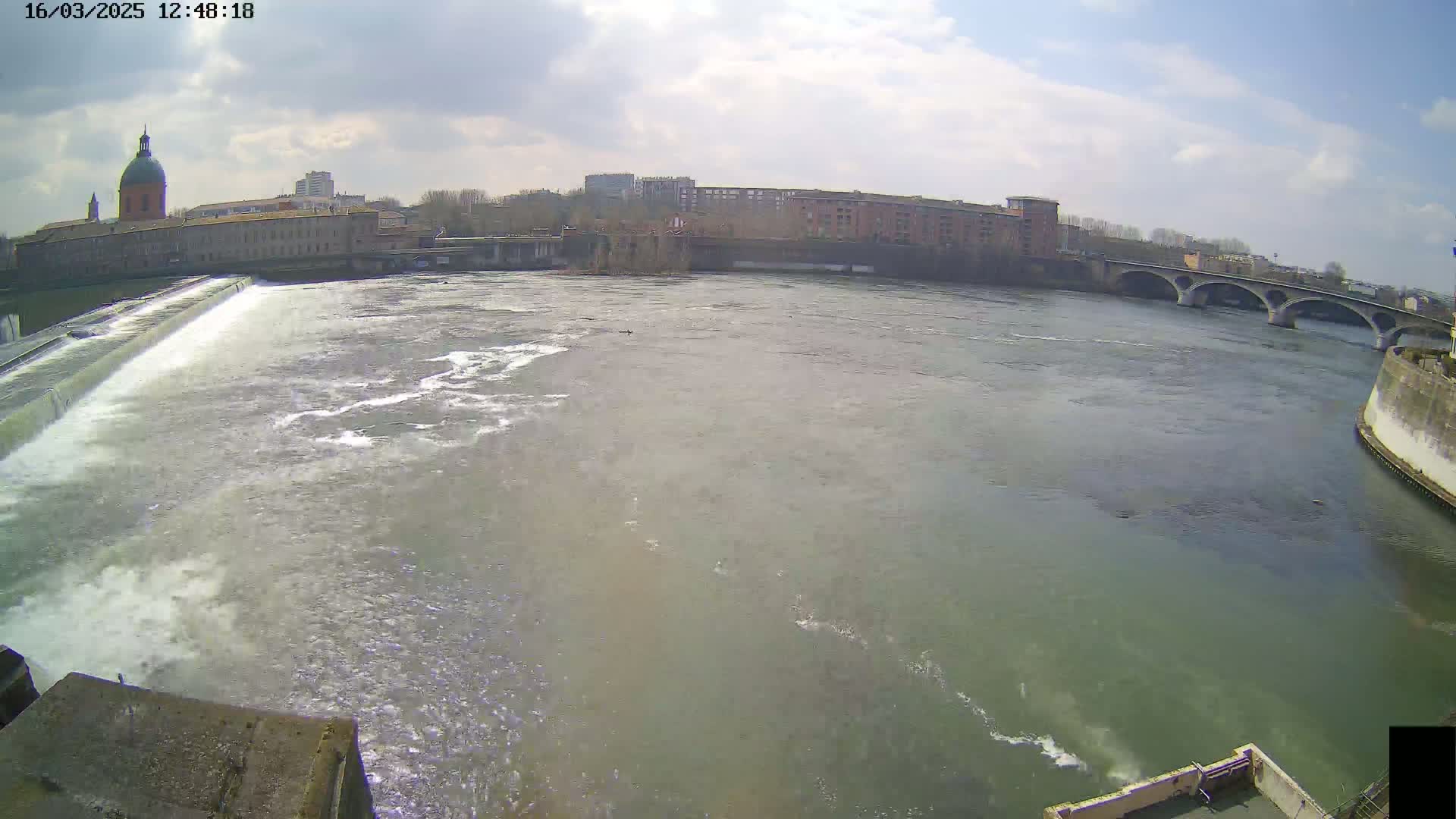 A wide shot of a river flowing past a dam, with a city and a bridge visible in the background under partly cloudy skies.
