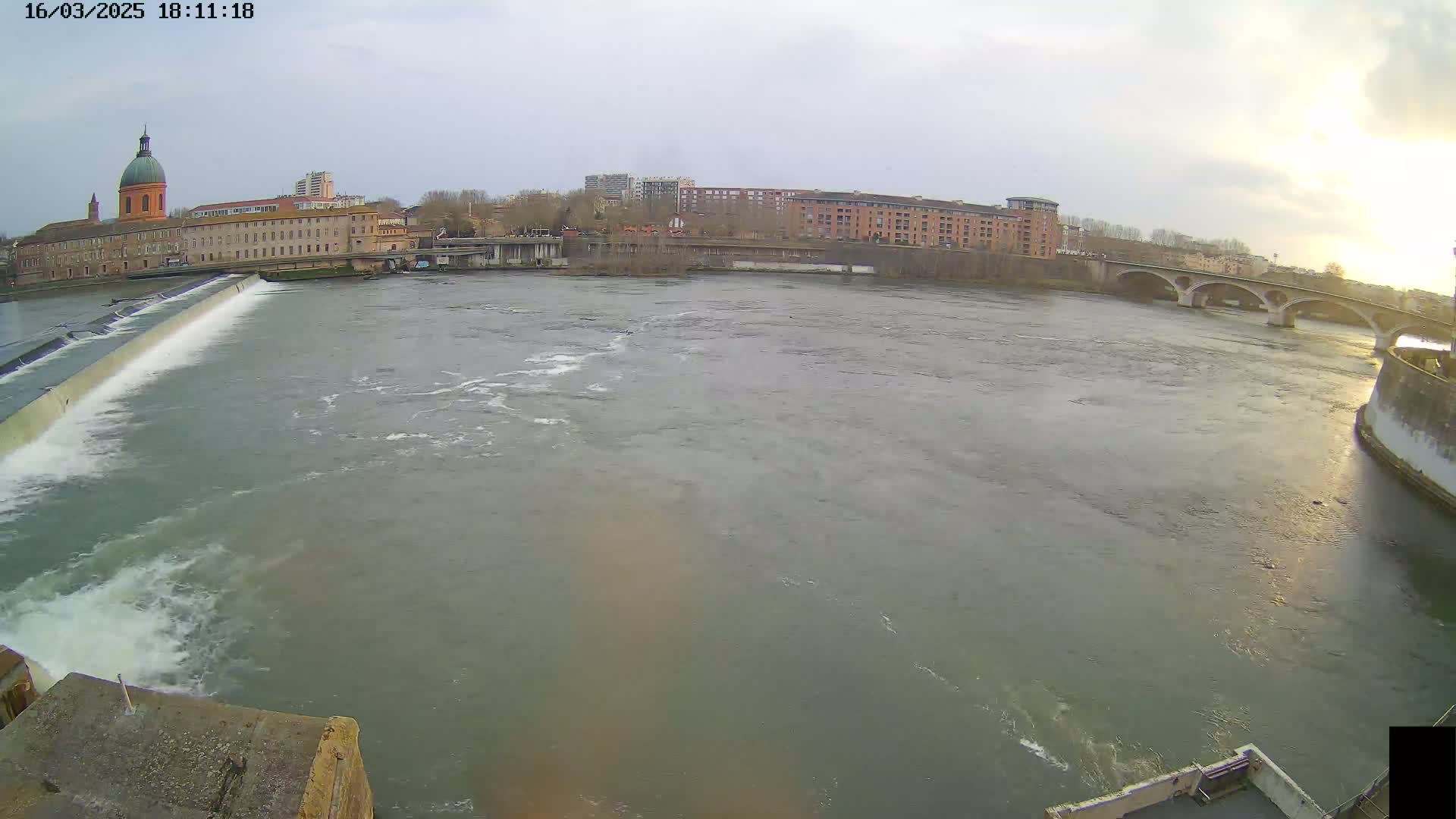 A wide shot of a river flowing past a dam and under a bridge, with buildings and a domed structure visible on the far bank under a cloudy sky.