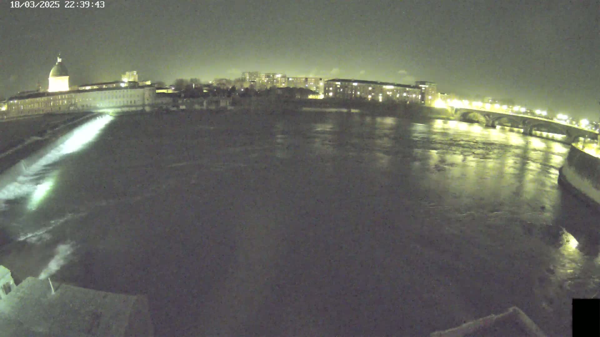 A nighttime view of a river flowing past a city with buildings and a bridge, under a dark sky.