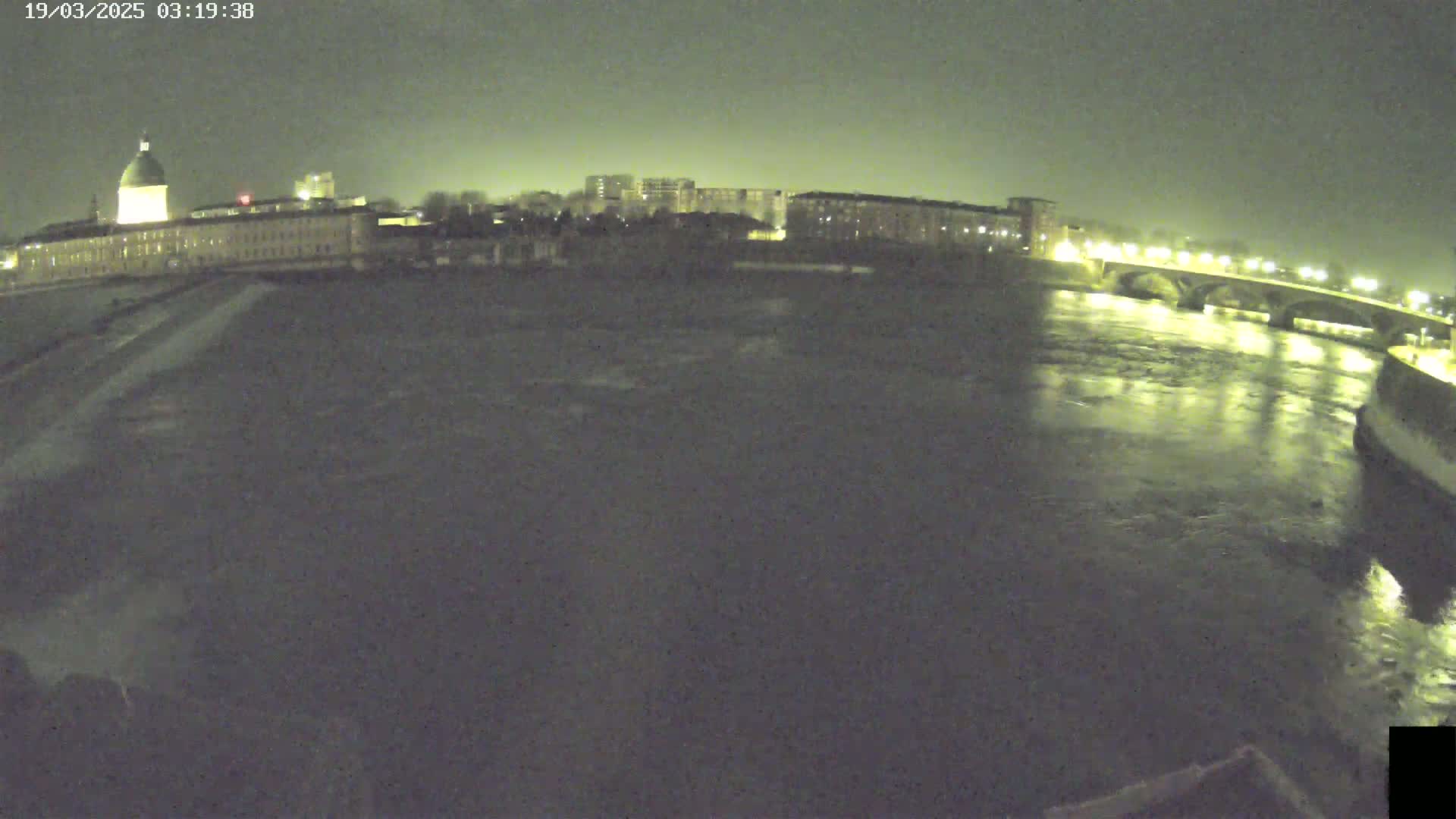 A nighttime view of a river flowing past city buildings and a bridge under a dark sky.