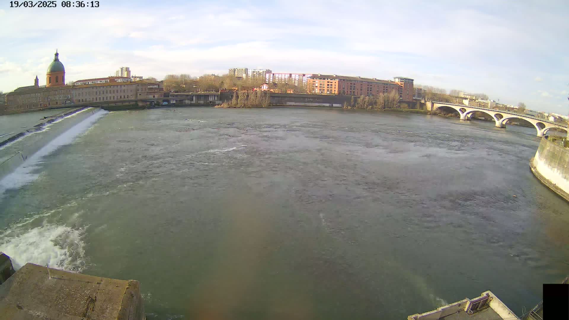 A wide shot of a river flowing under a bridge on a partly cloudy day, with buildings and a dome-topped structure visible on the far bank.