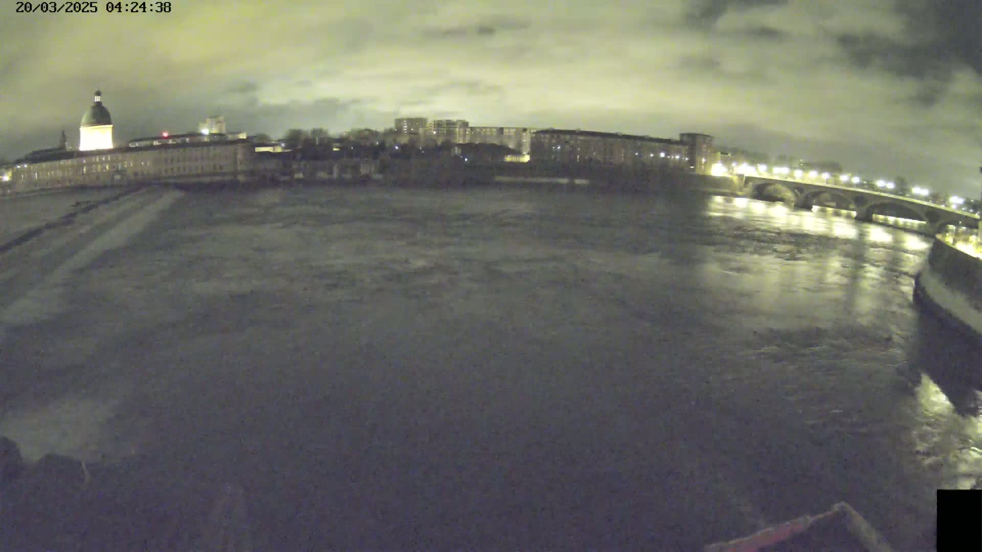 A wide nighttime shot shows a river flowing past city buildings and a bridge under a cloudy sky.