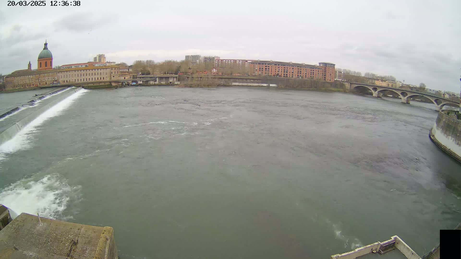 A wide shot shows a river flowing past a dam and under a bridge, with buildings and a domed structure visible in the background under an overcast sky.