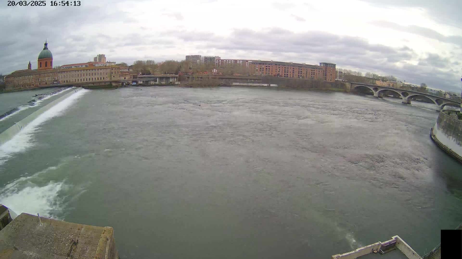 A wide shot of a river flowing under an overcast sky past a dam, buildings, and a bridge.