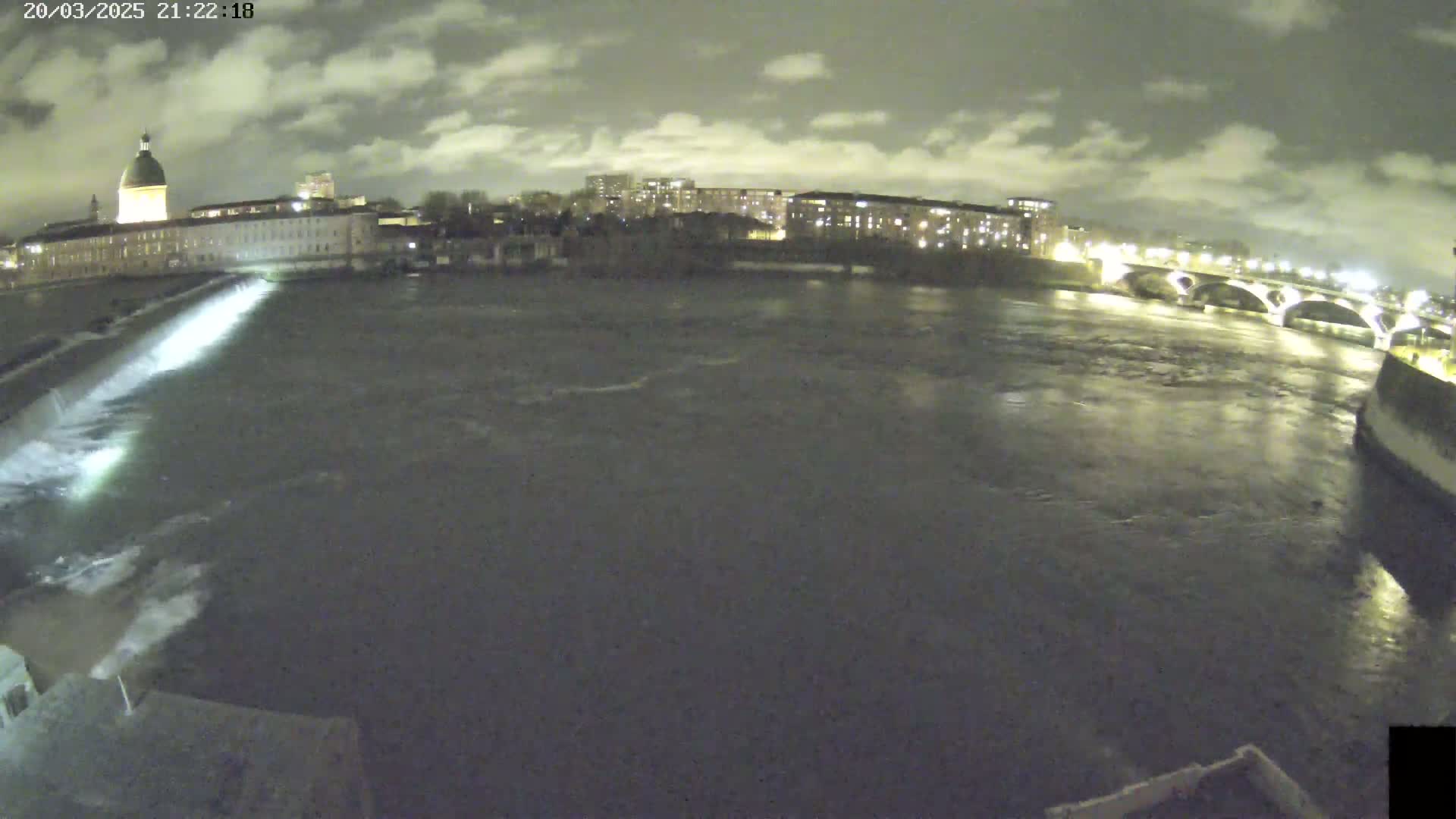 A nighttime view of a river flowing past city buildings and a bridge under a cloudy sky.