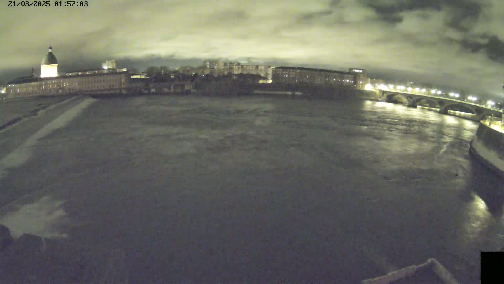 A wide nighttime shot shows a dark river flowing under a bridge past a large building with a dome under a cloudy sky.