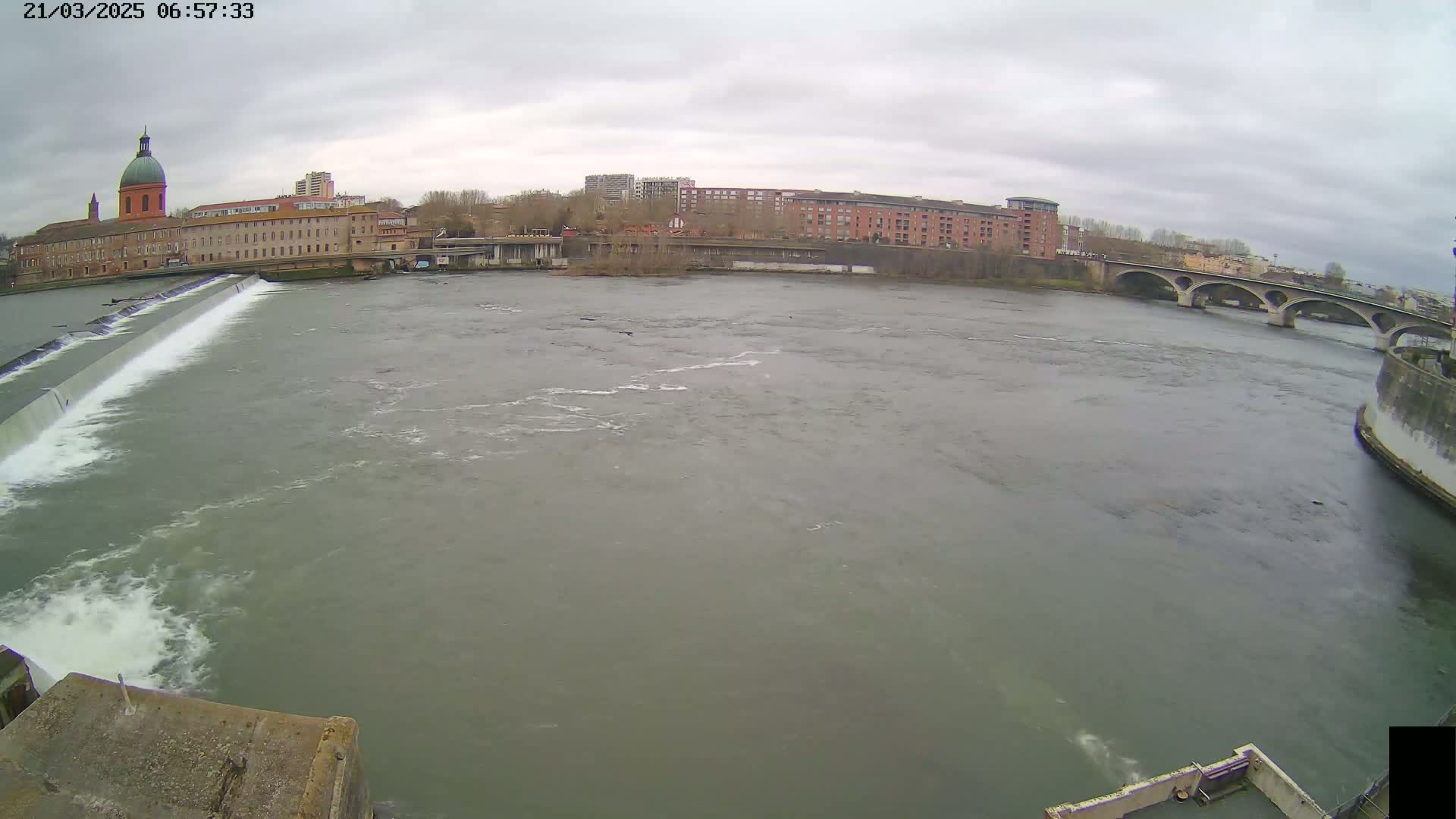 A wide shot of a river flowing under a bridge on an overcast day, with a dam and buildings visible in the background.