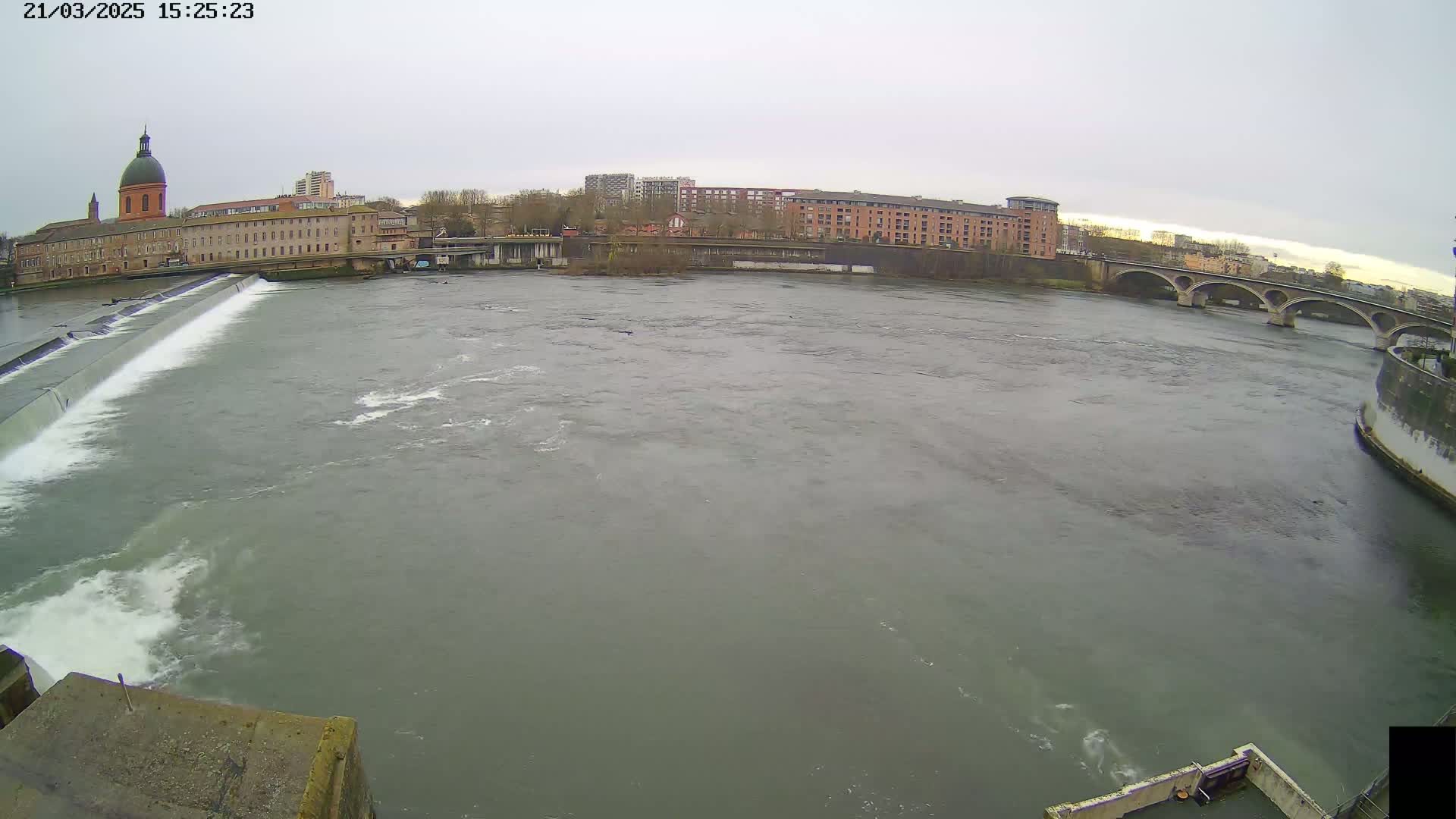 A wide, grey river flows past a city with buildings and a stone bridge under an overcast sky.
