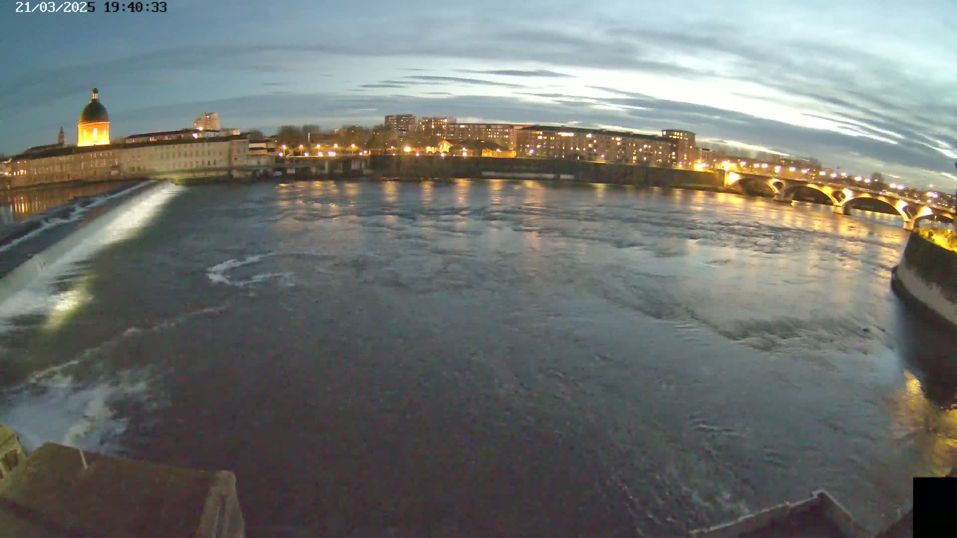 A wide shot at twilight shows a river flowing past a city with buildings lit up at night, a dam, and a bridge.