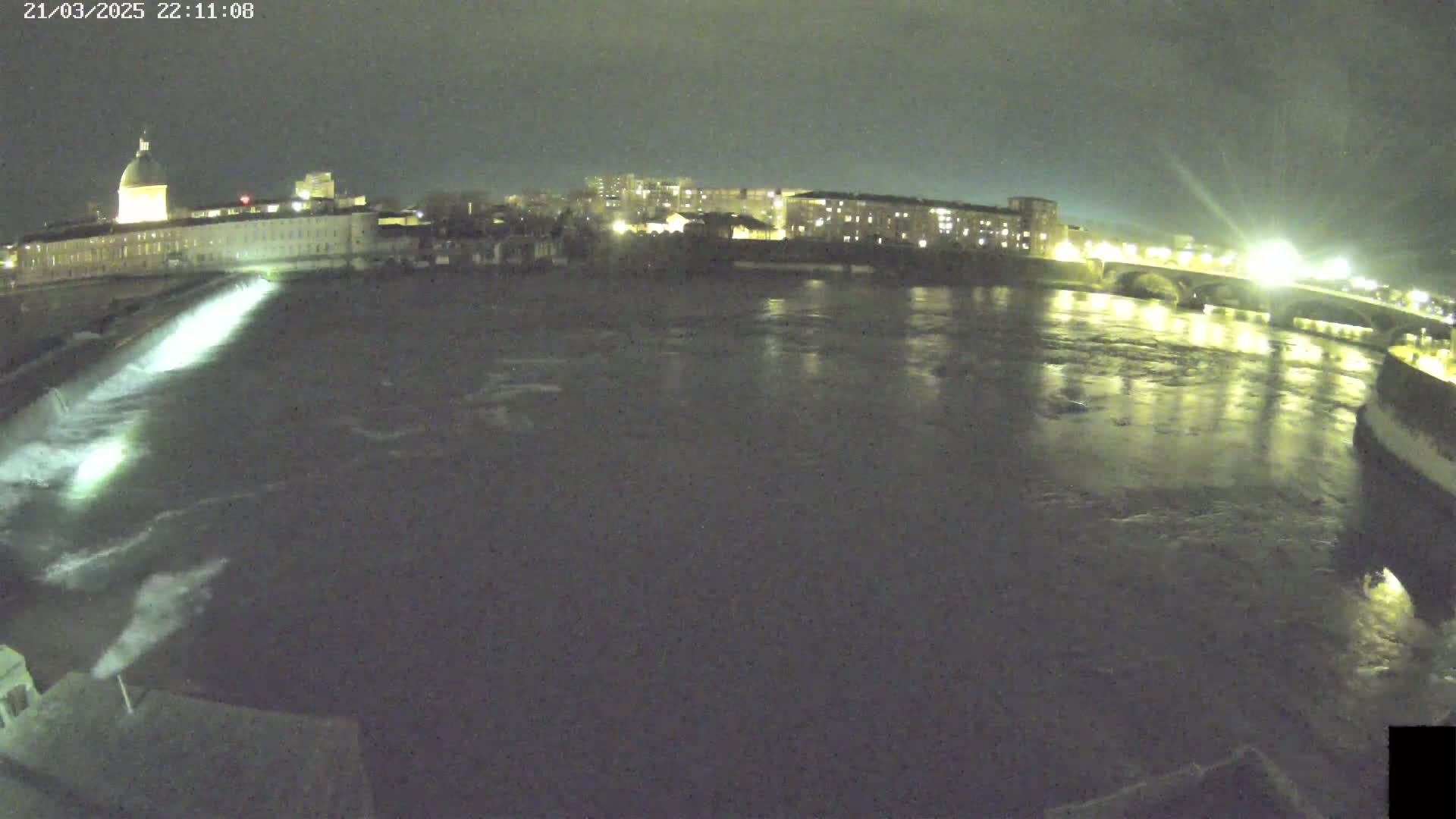 A nighttime view of a river flowing under a bridge past buildings, with a structure causing water to rush over a dam.