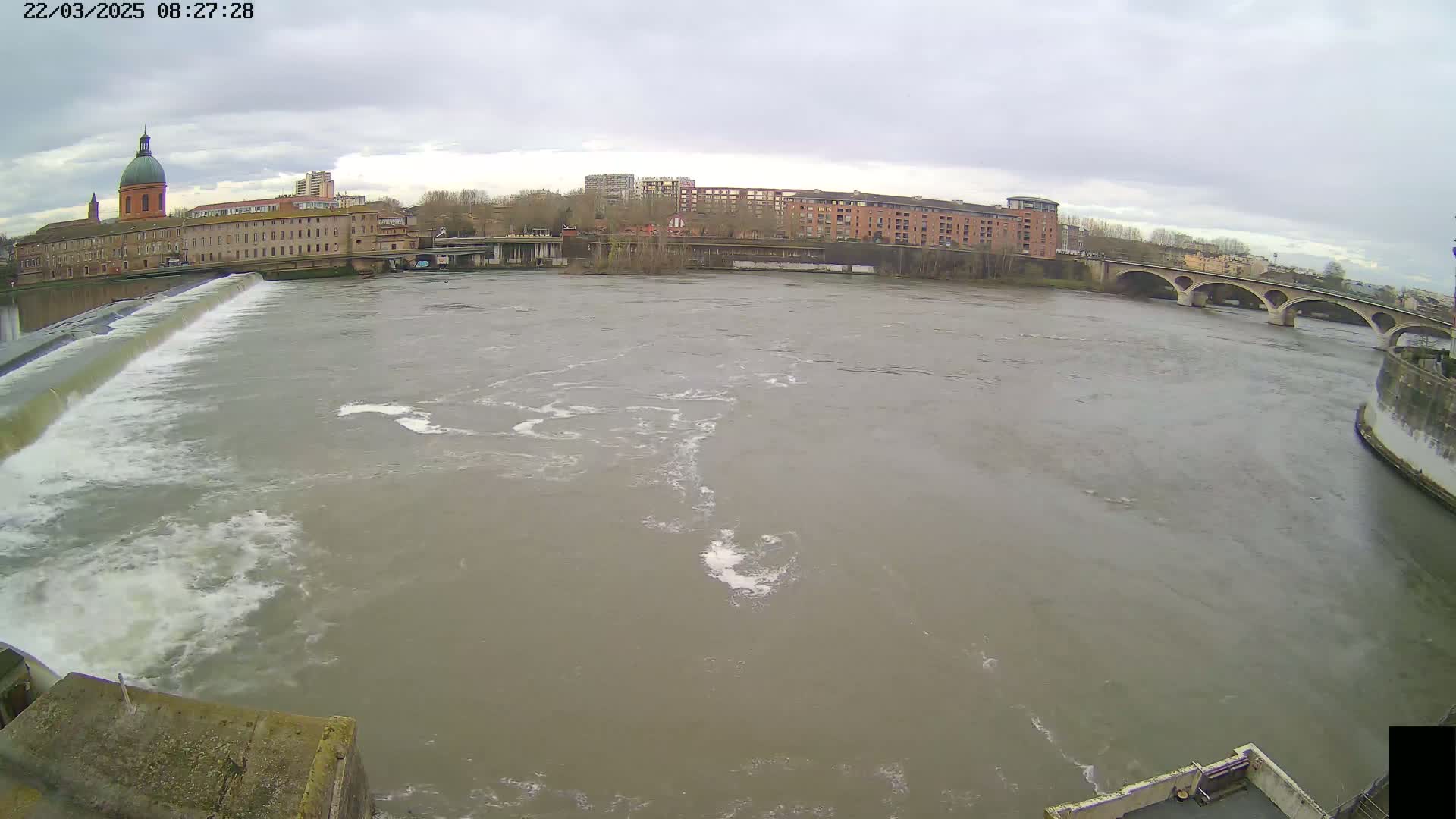 A wide shot of a river flowing past a city under an overcast sky, with a dam visible in the foreground and a bridge in the background.