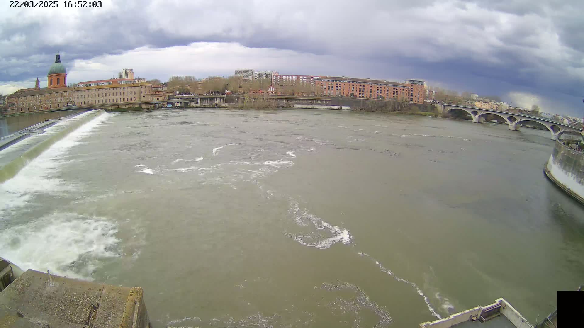 A wide shot shows a river flowing under an overcast sky past a dam and buildings, with a bridge in the background.