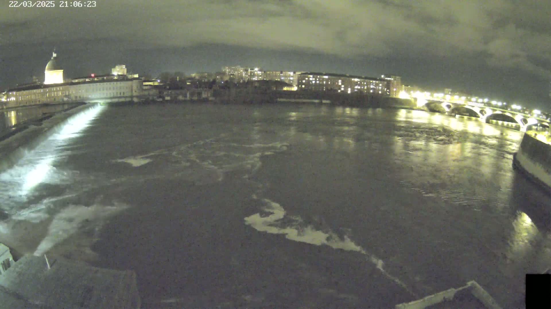 A nighttime view of a river flowing around a city, with a dam and bridge illuminated under a cloudy sky.