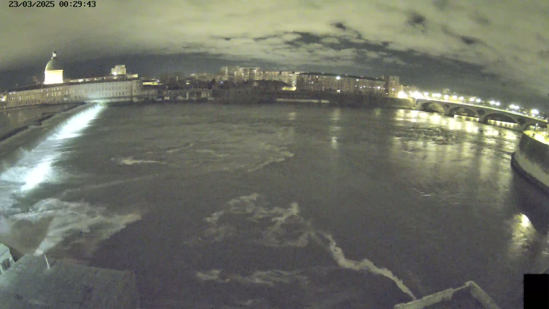 A nighttime view of a river flowing past a city with a dam releasing water, under a cloudy sky.