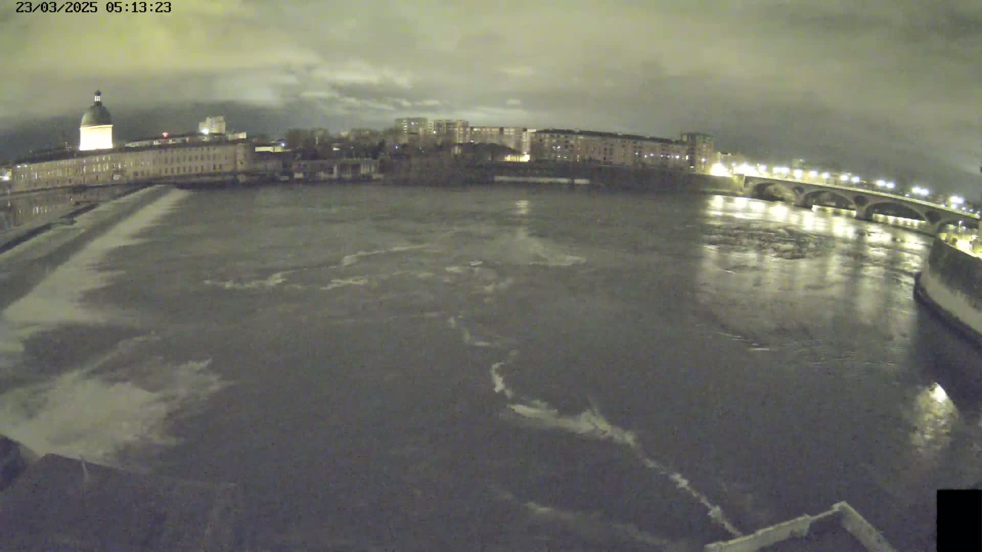 A nighttime view of a river flowing past city buildings and a bridge under a cloudy sky.
