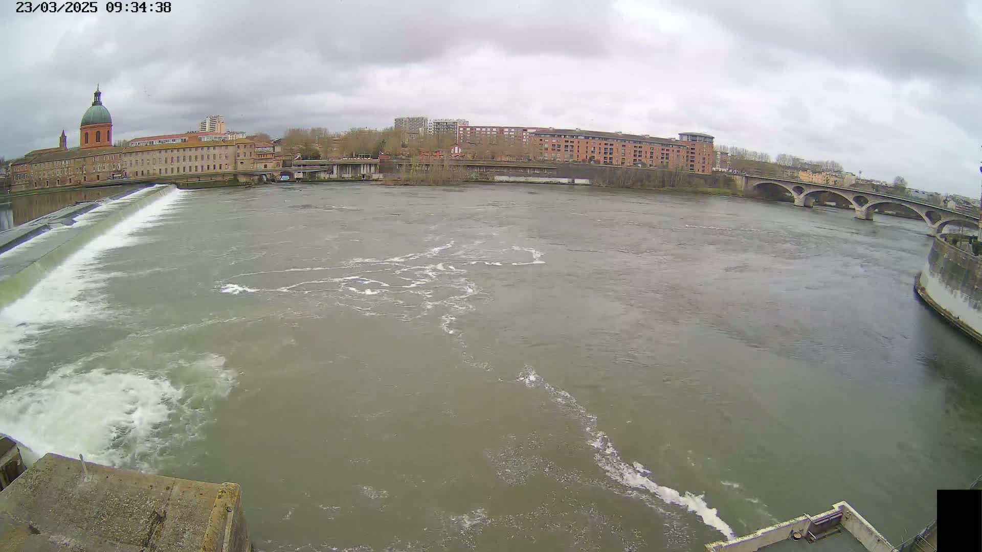 A wide, murky river flows under an overcast sky past a dam and buildings, including a large building with a dome.