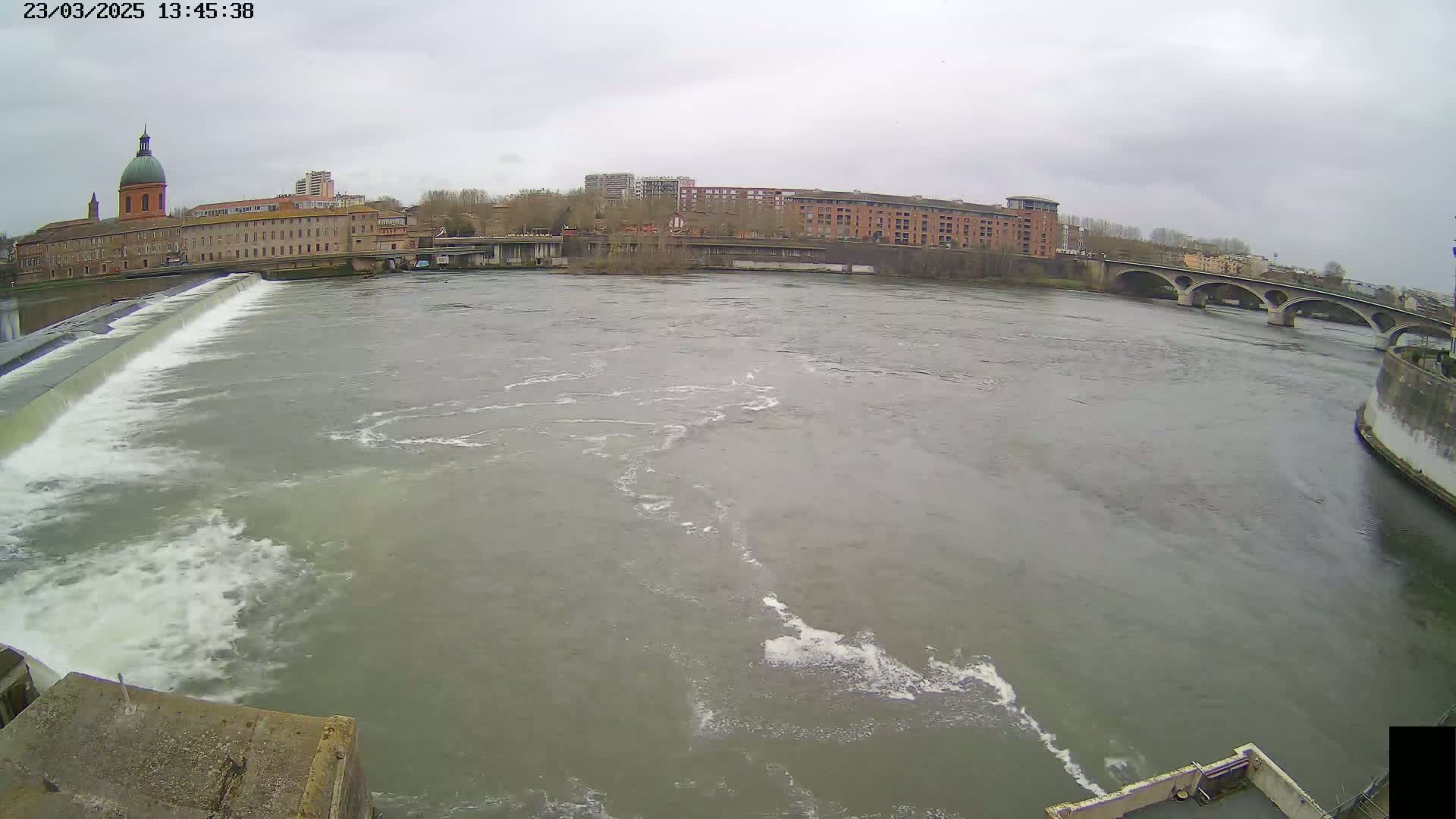 A wide river flows past a city with buildings and a bridge under an overcast sky.