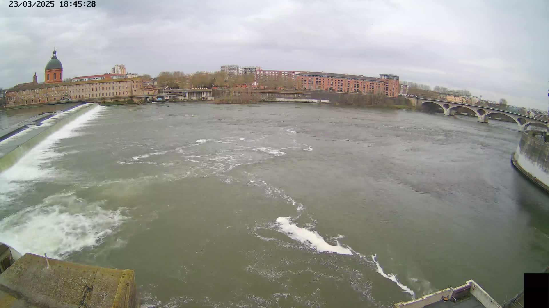 A wide shot of a river flowing over a dam under an overcast sky, with buildings and a bridge visible in the background.