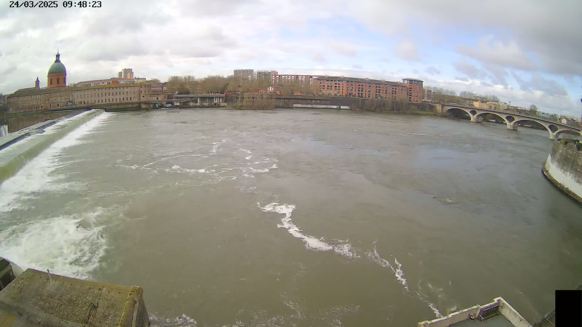 A wide shot of a river flowing under an overcast sky past a dam and buildings, with a bridge visible in the distance.