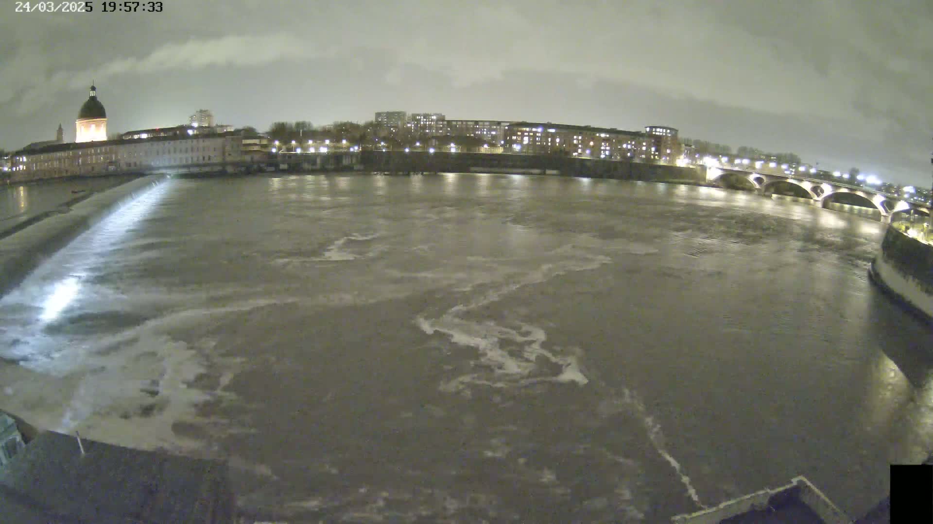A nighttime view of a river flowing past city buildings under a cloudy sky, with a dam releasing water into the river.