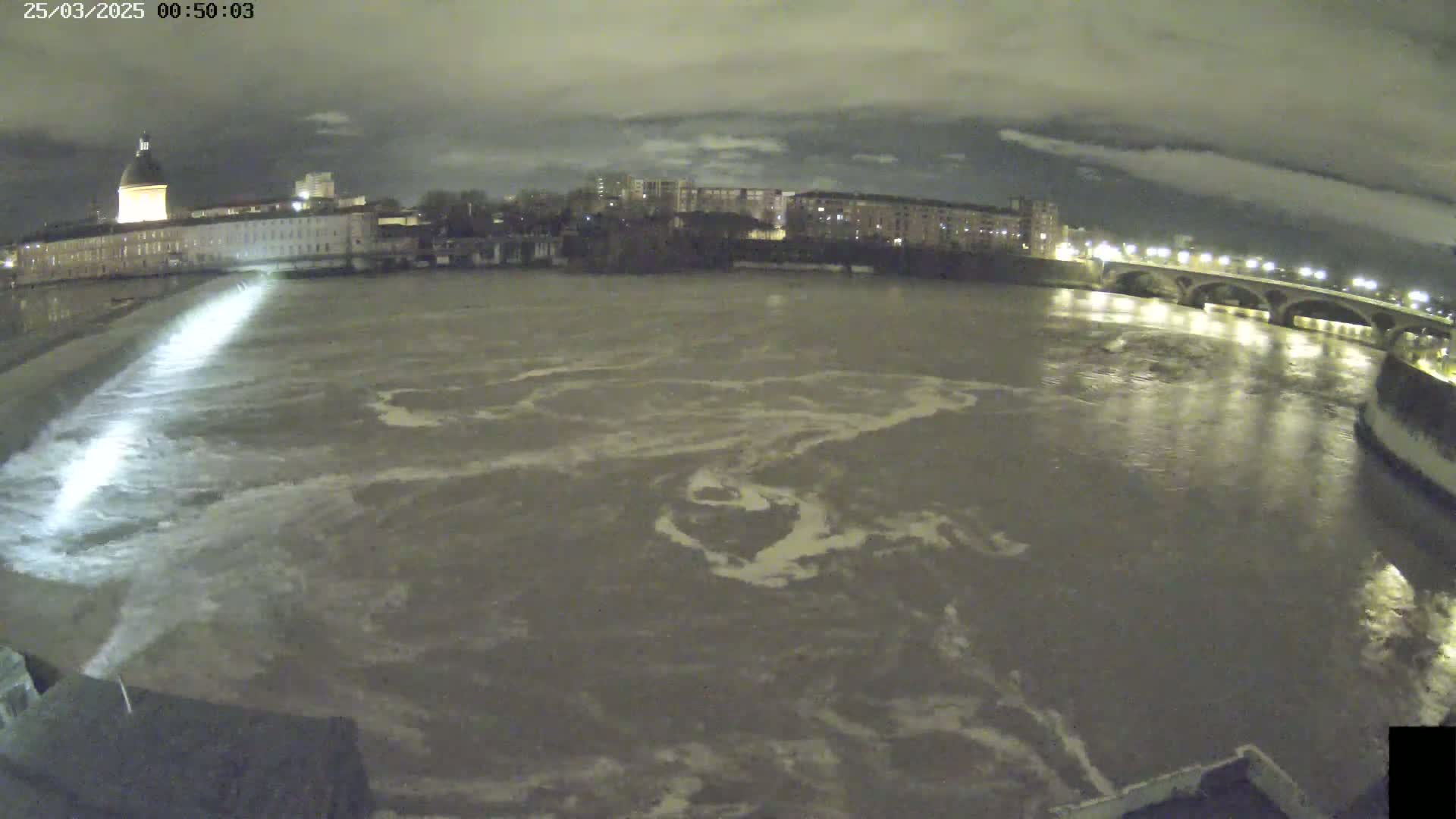 A nighttime view of a wide, murky river flowing past city buildings and a bridge under a cloudy sky.