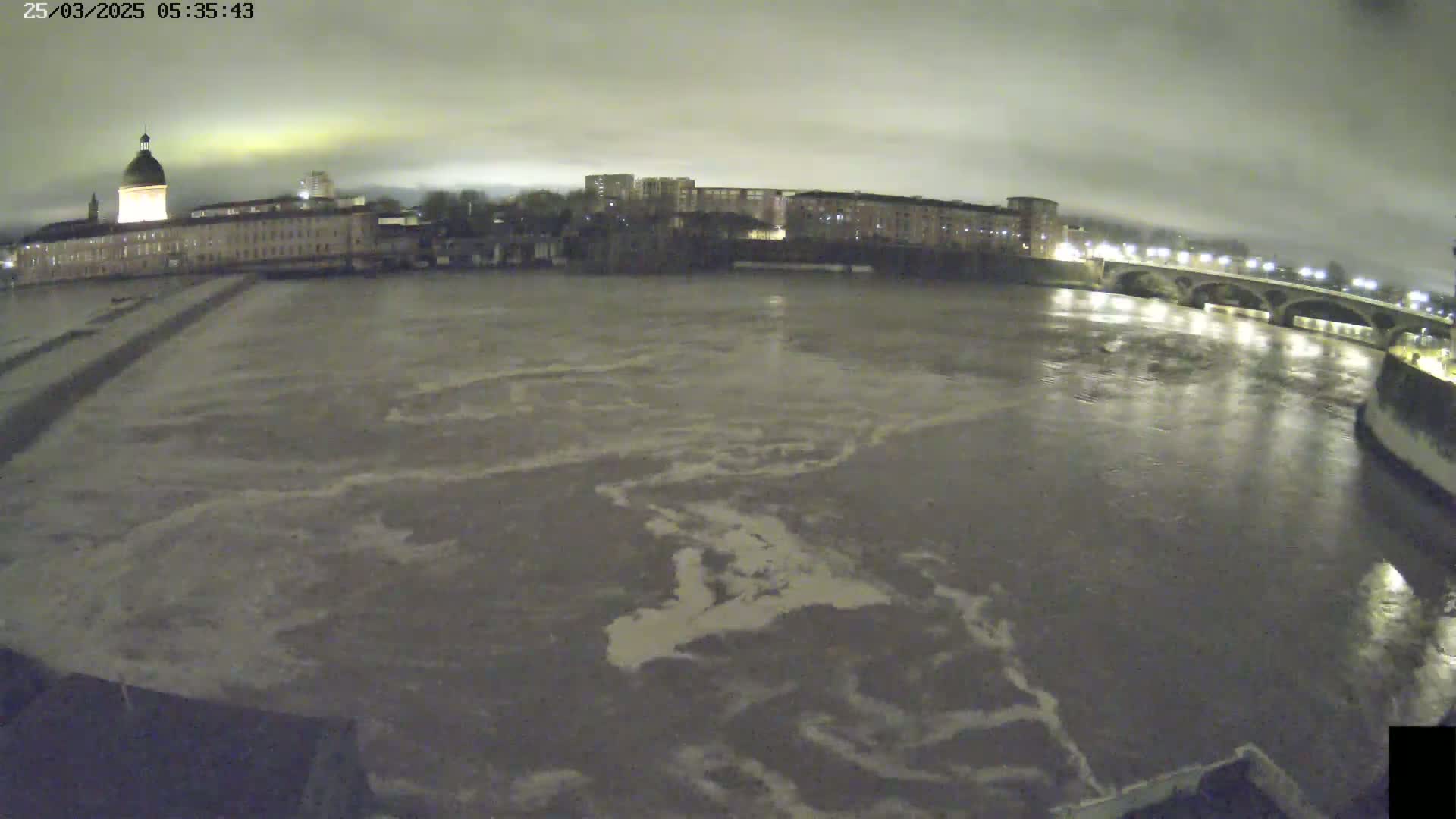 A wide shot at night shows a river flowing rapidly past buildings and a bridge under an overcast sky.