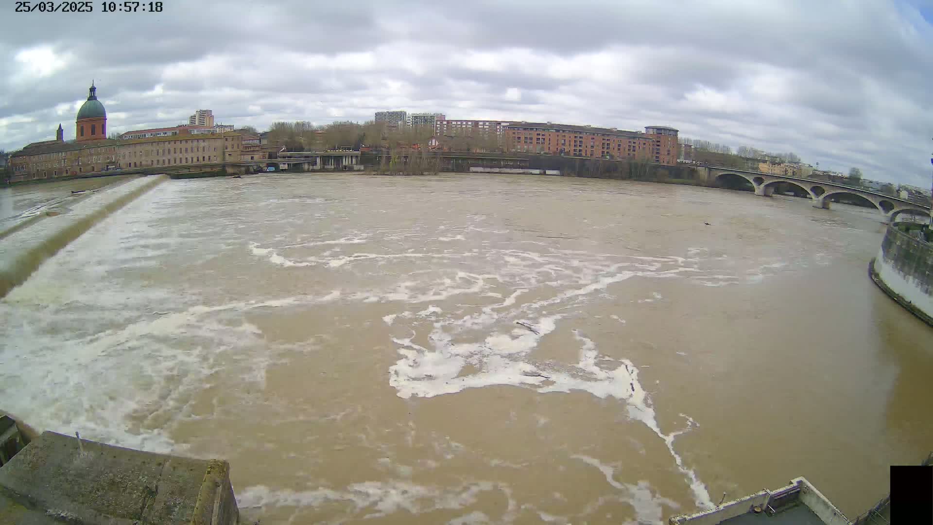 A wide, muddy river flows rapidly under an overcast sky past a city with buildings and a bridge.