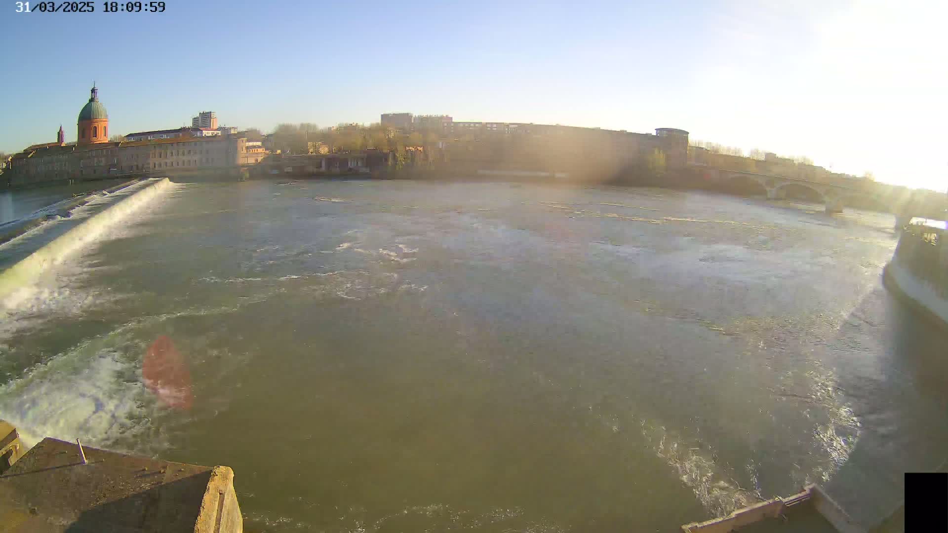 A wide shot shows a river flowing past a city with a dam and bridge under a mostly clear sky.