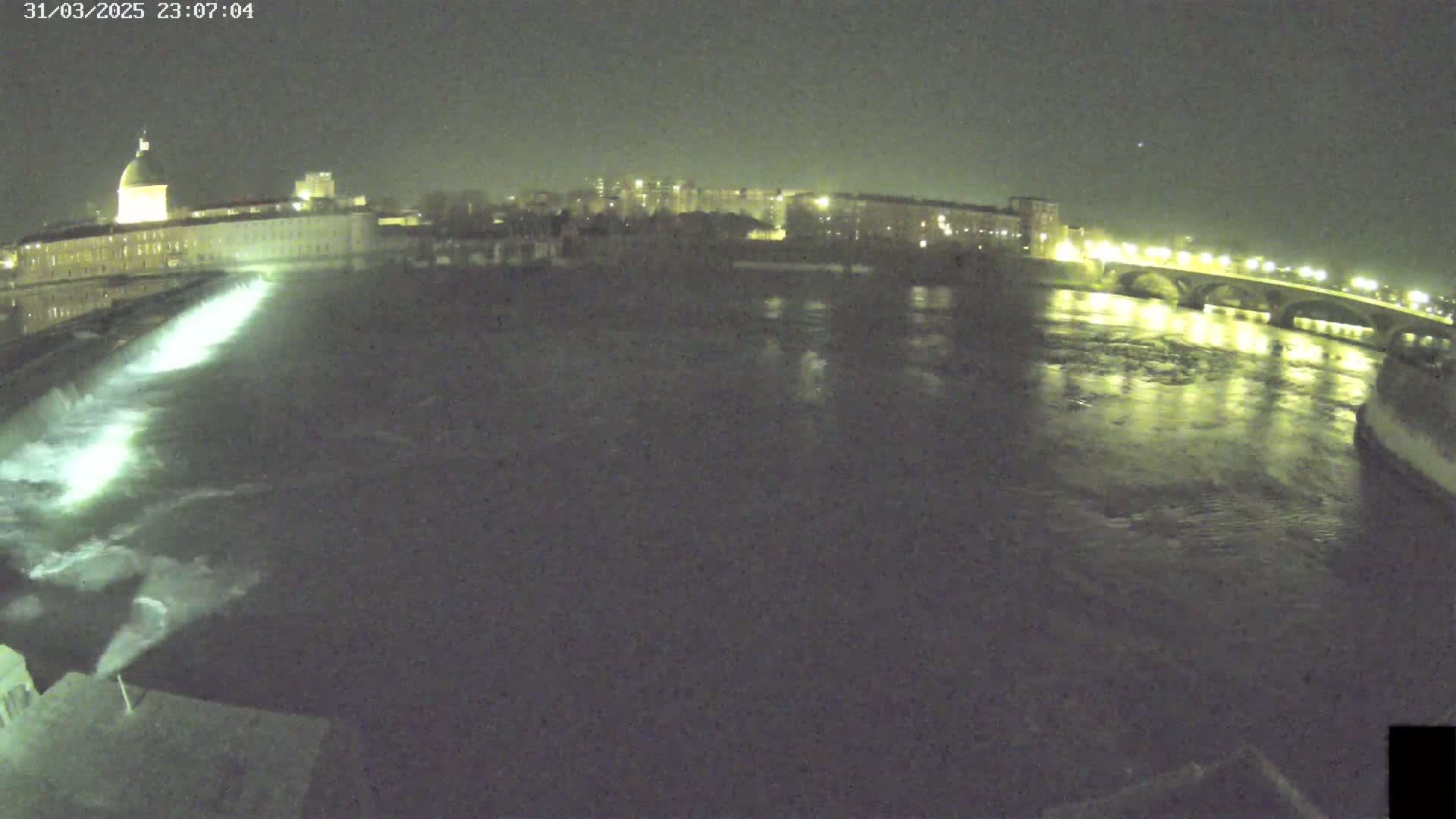 A nighttime view of a river flowing past a city, with a dam releasing water and a bridge visible in the distance under a clear, dark sky.
