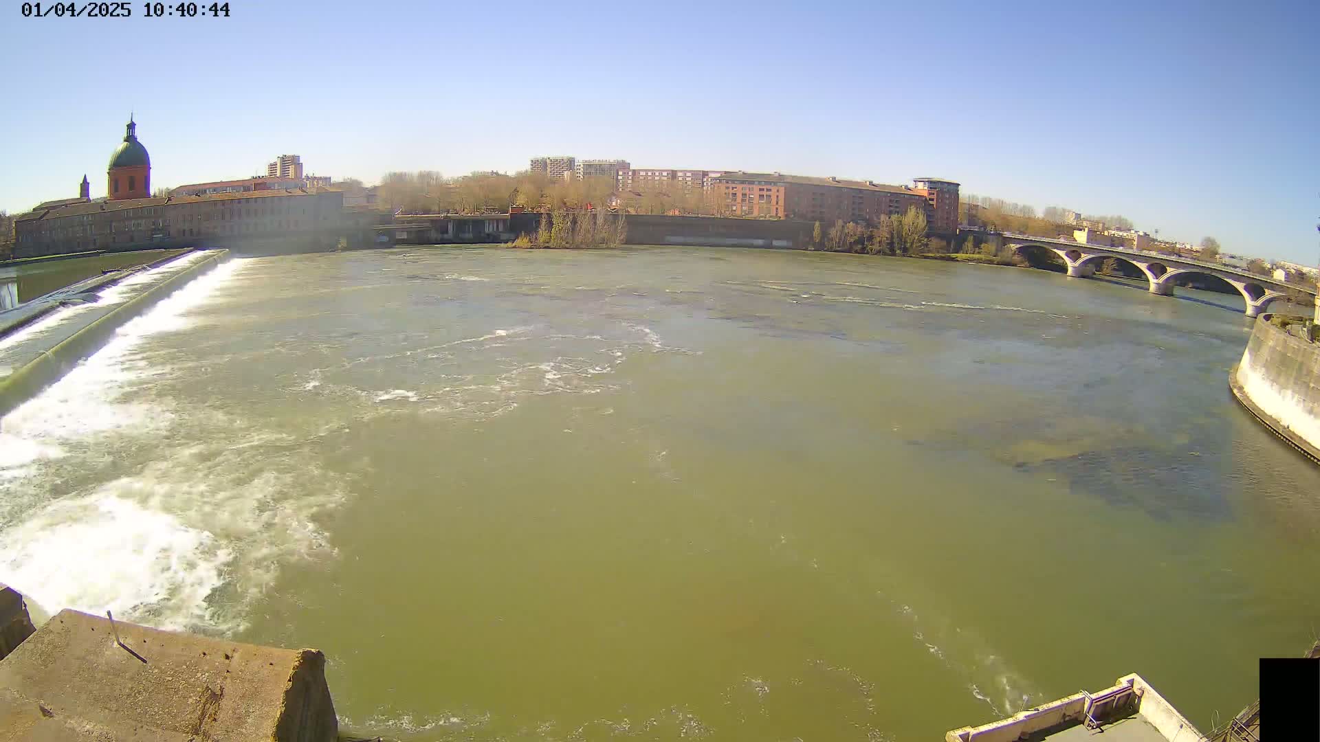 A wide shot of a river flowing under a bridge on a sunny day, with a dam and buildings visible in the background.