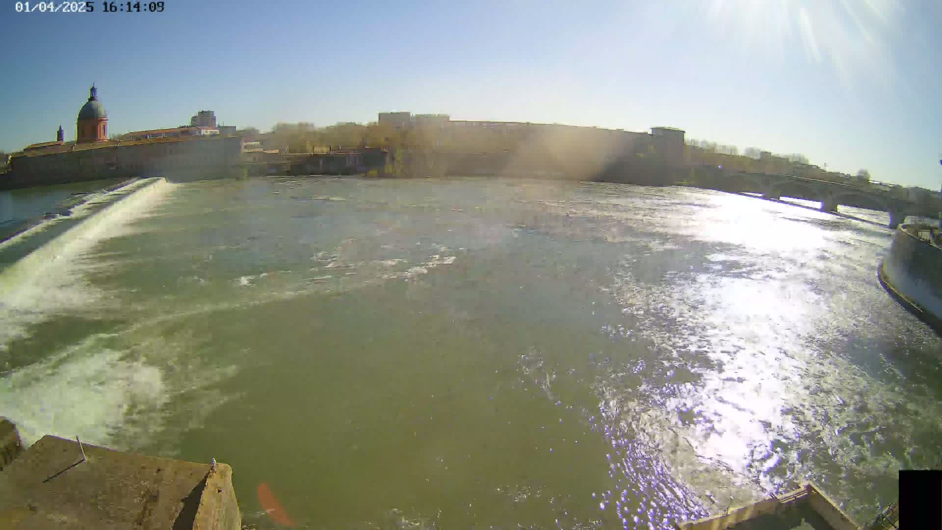 A wide shot shows a river flowing past a dam and under a bridge on a sunny day, with buildings and a dome-topped structure visible in the background.