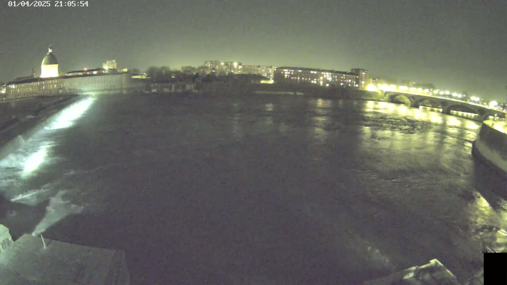 A nighttime view of a river flowing past a city, with a dam releasing water and lights illuminating the cityscape and bridge.