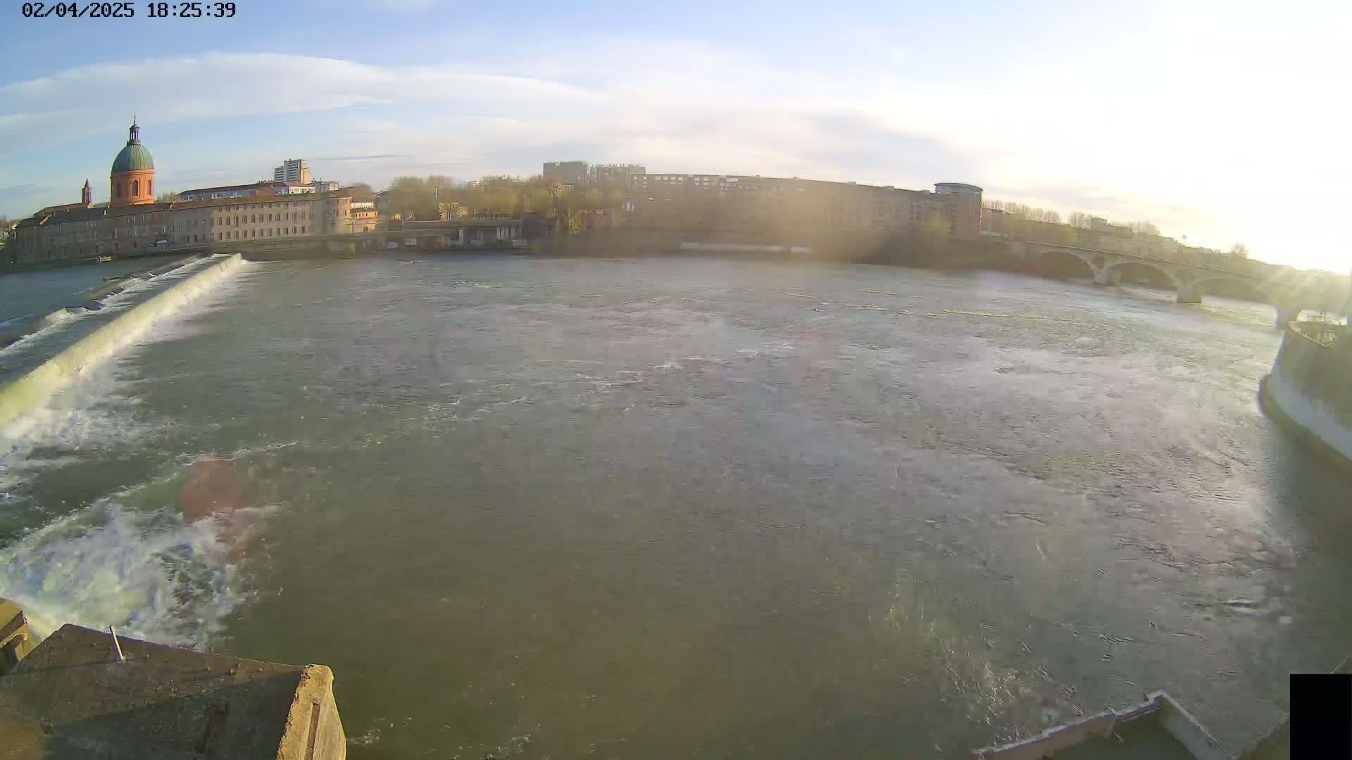 A wide shot of a river flowing past a dam, with a city skyline and a domed building visible in the background under a mostly clear sky.